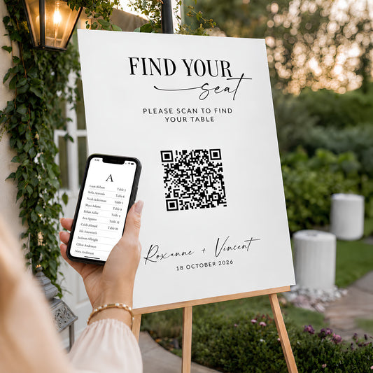 A wedding reception seating sign on an easel in an outdoor garden setting, with a hand holding a smartphone showing the mobile seating app next to the printed sign.