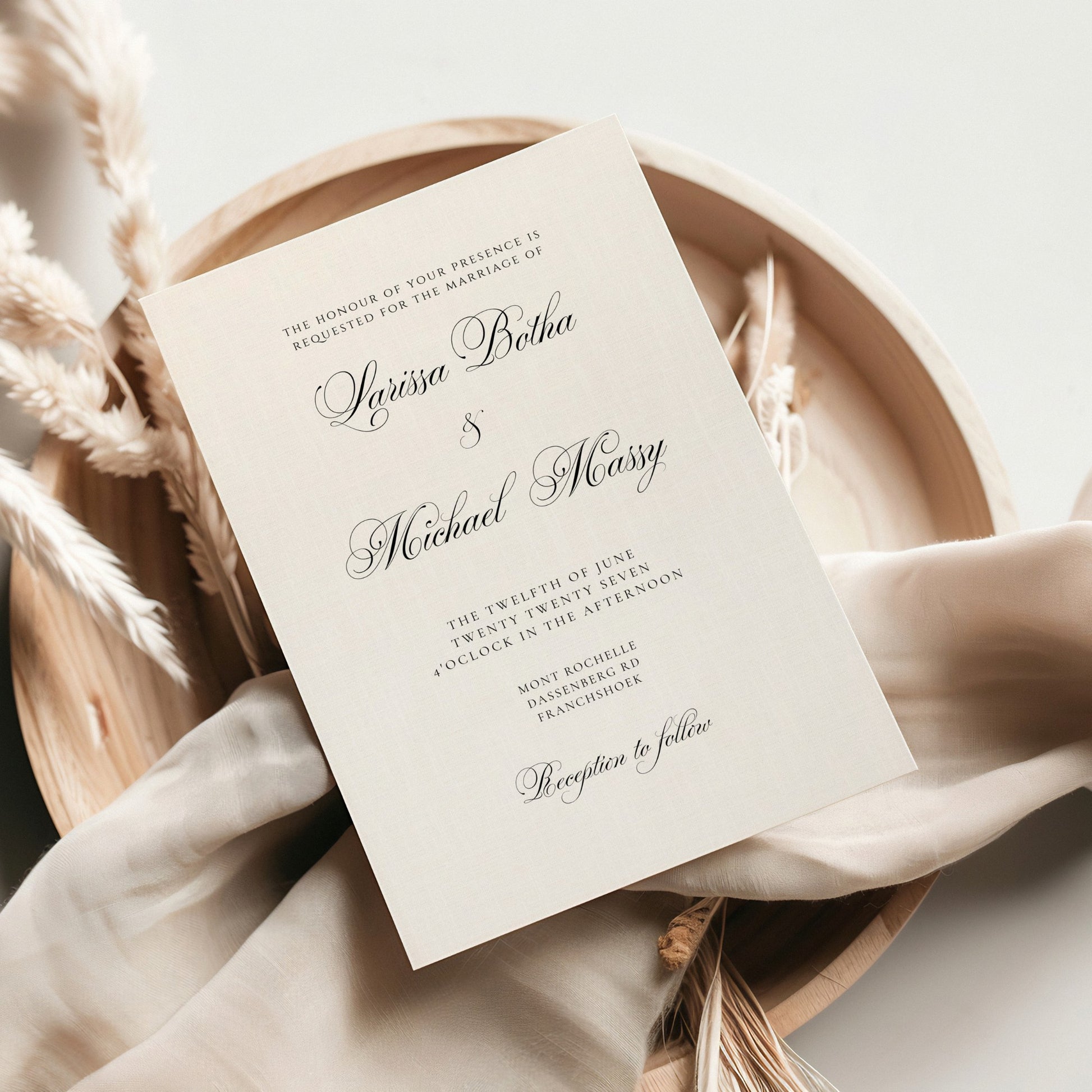 A wedding invitation card with elegant black script text on an off-white paper background, styled on top of a light beige fabric and dried pampas grass inside a round wooden tray.