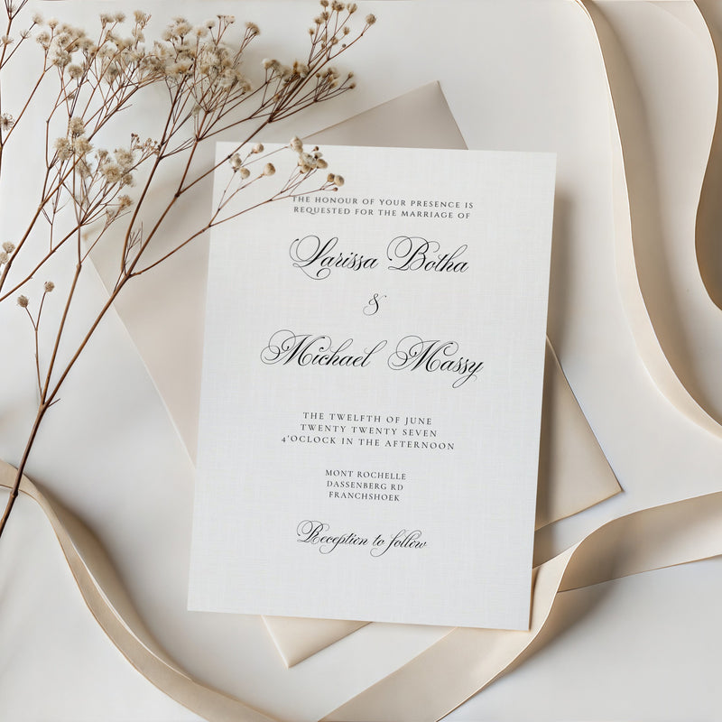 A white wedding invitation card with elegant black script text, layered over a cream envelope and surrounded by cream ribbon and dried baby's breath flowers on a light neutral background.