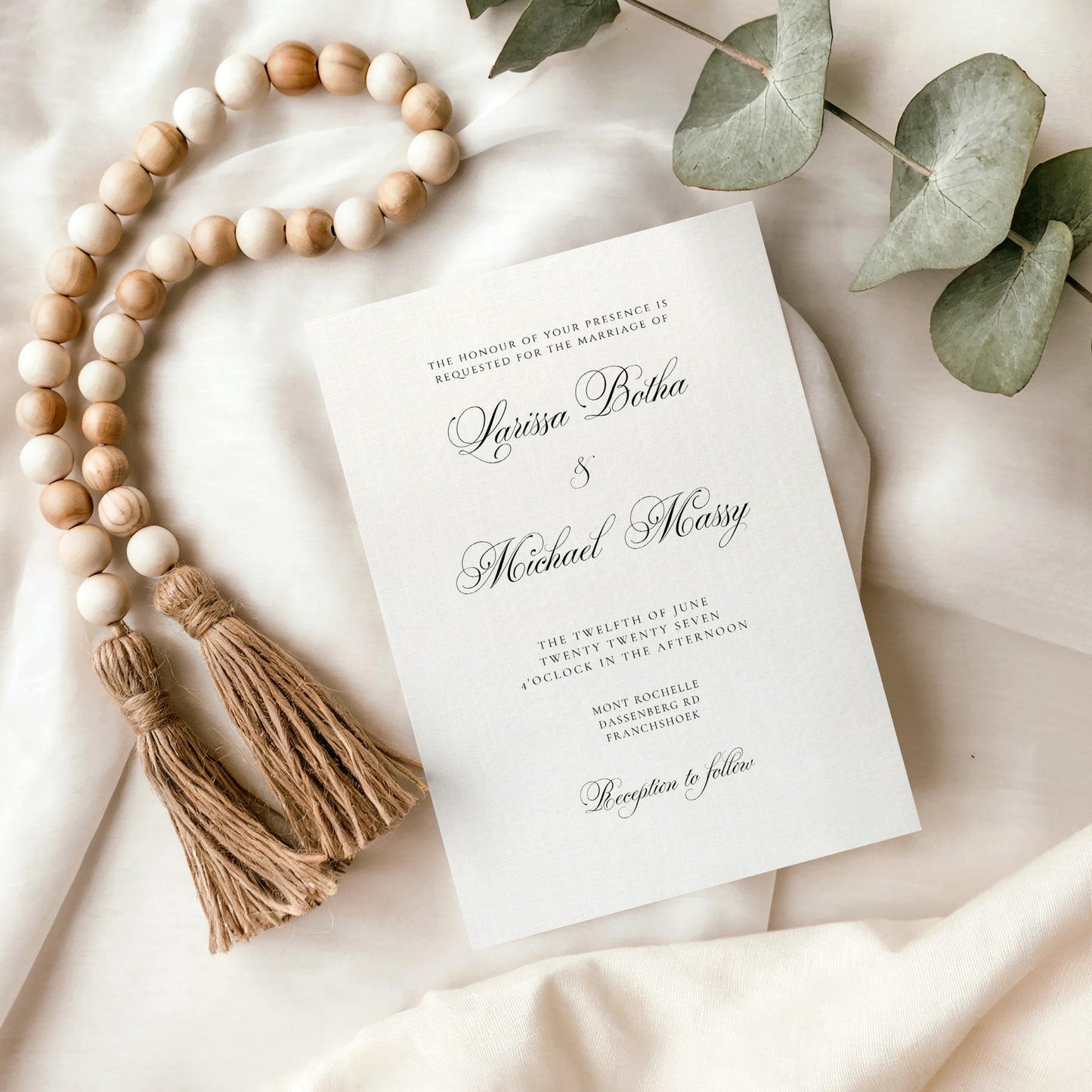 A wedding invitation card with elegant black script text on a white background, styled in a flat lay with a wooden bead garland, tassels, and green eucalyptus leaves on draped cream fabric.