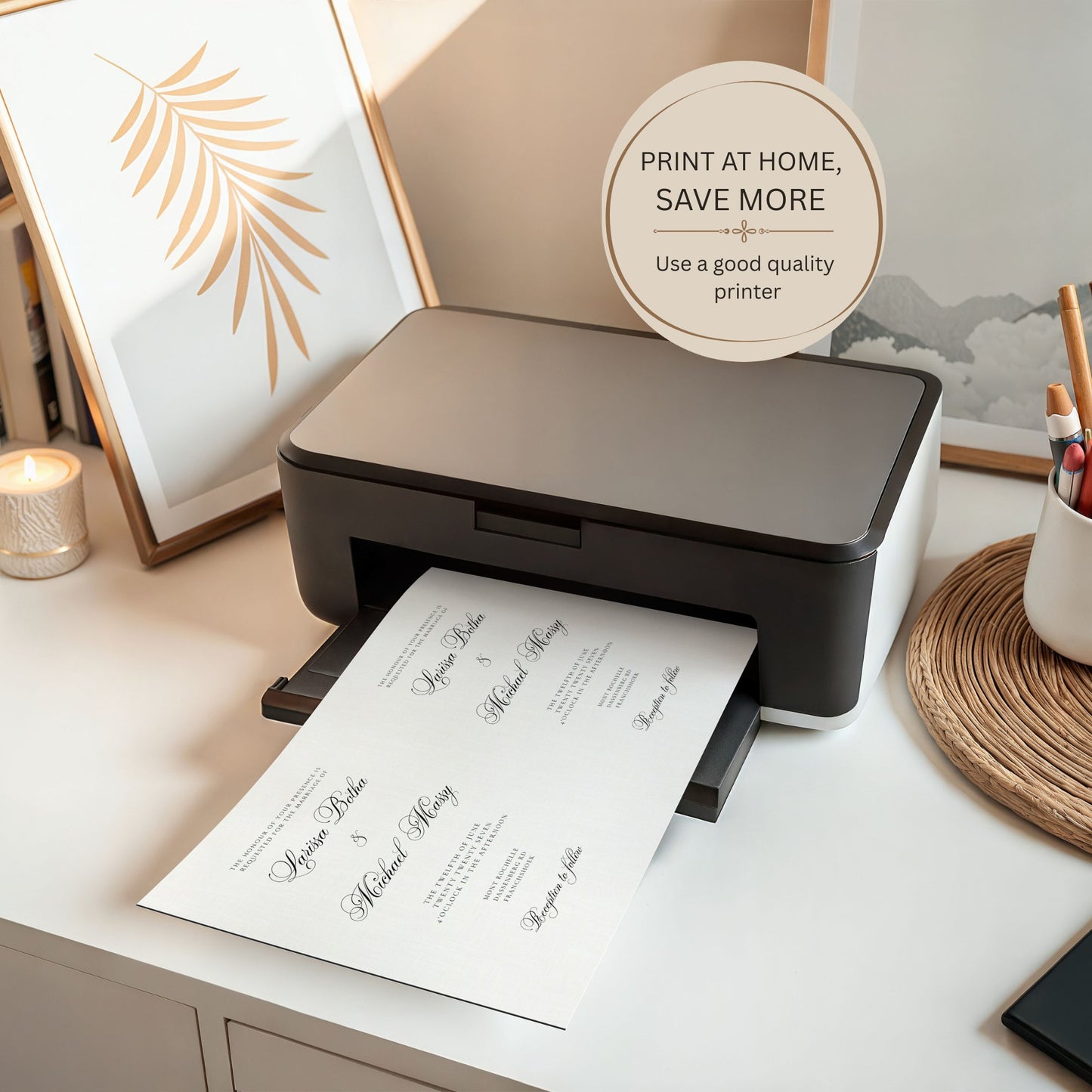 A compact black and white home printer sits on a white desk, printing a wedding invitation card. The scene is styled with a gold-framed leaf print, a lit candle, and stationery, with a circular badge reading 'PRINT AT HOME, SAVE MORE'.