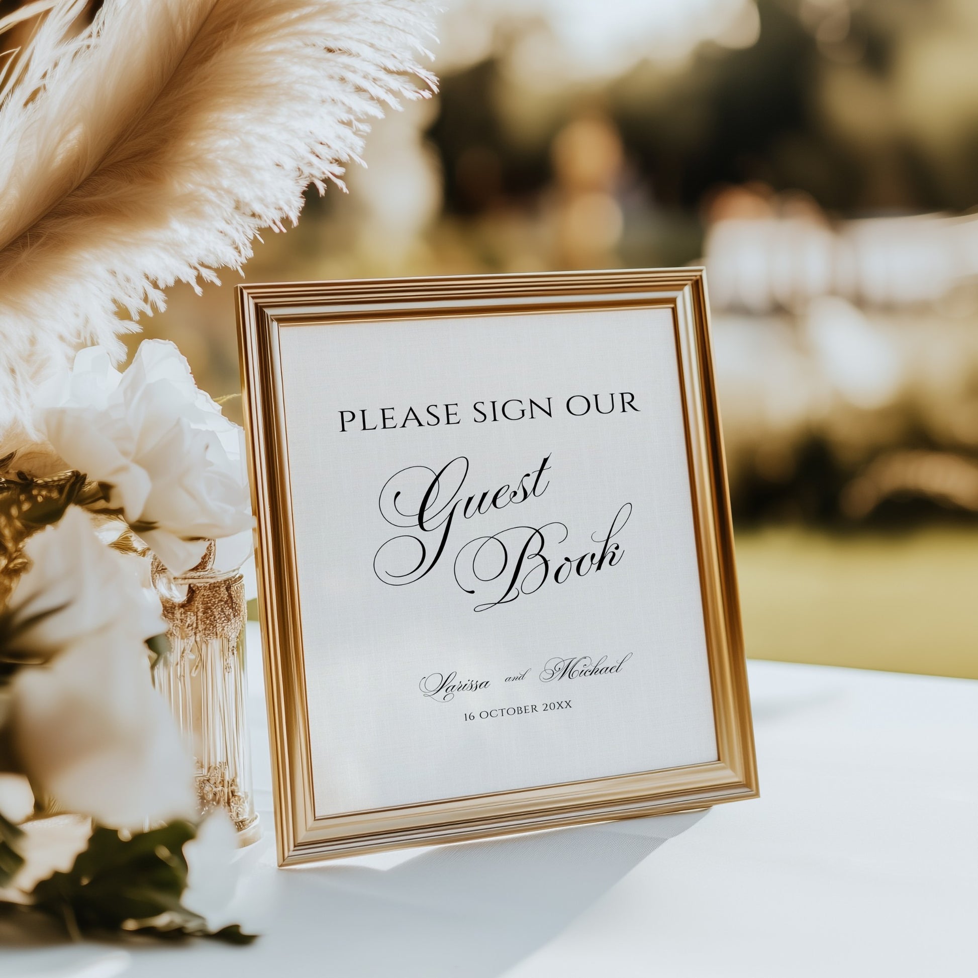 A gold-framed guest book sign with elegant black script text, displayed on a white table with decorative white flowers 